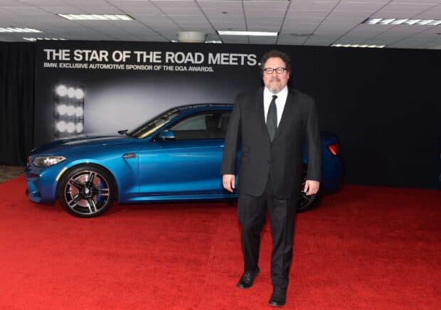 Jon Favreau stands confidently on a red carpet with a blue car behind him, HD Desktop Wallpaper