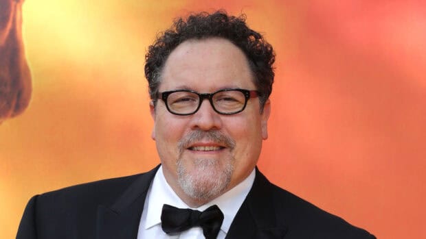 Jon Favreau is wearing a black tuxedo and glasses with a subtle smile in front of a warm background, HD Desktop Wallpaper