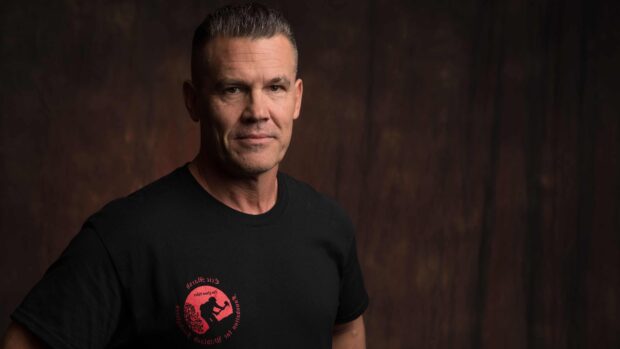 Josh Brolin stands wearing a black shirt with a red logo on dark background, 4K Desktop Wallpaper