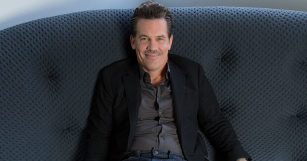 Josh Brolin sitting relaxed on a dark textured sofa smiling in a black jacket, 2K Desktop Wallpaper