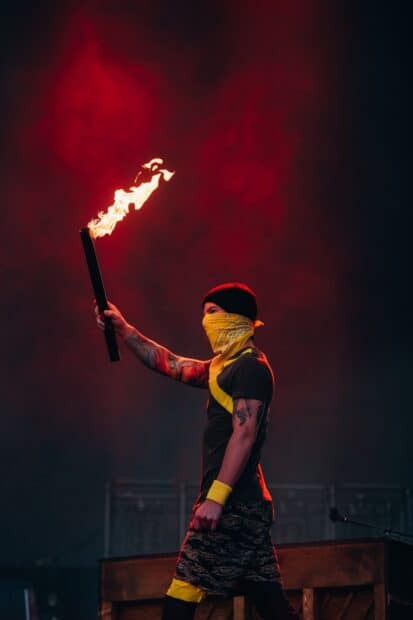Josh Dun is holding a flaming torch while wearing a yellow bandana and black beanie on stage, HD Desktop Wallpaper