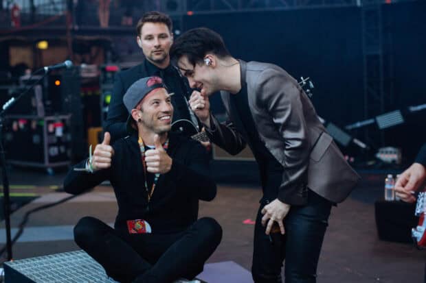 Josh Dun smiles while sitting and interacting with a man in a shiny jacket backstage, HD Desktop Wallpaper