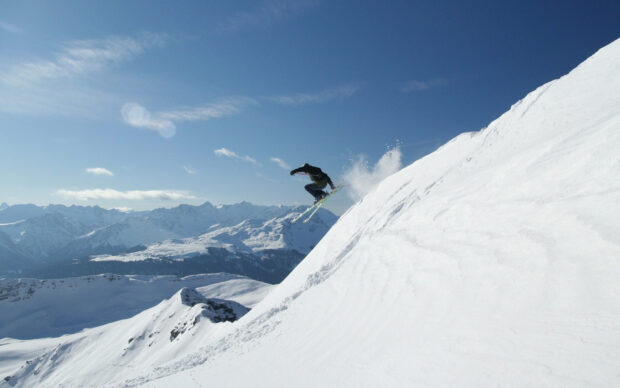 A skier is jumping off a snowy slope with mountains in the background showcasing skiing wallpapers, 2K Desktop Wallpaper