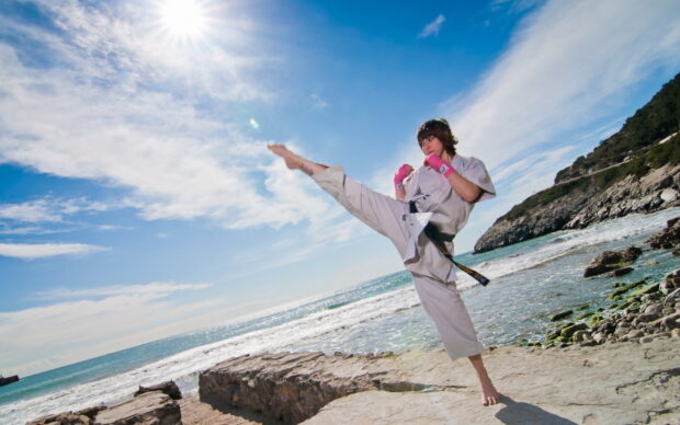 A person performing a high karate kick on rocky shore under bright blue sky, HD Desktop Wallpaper