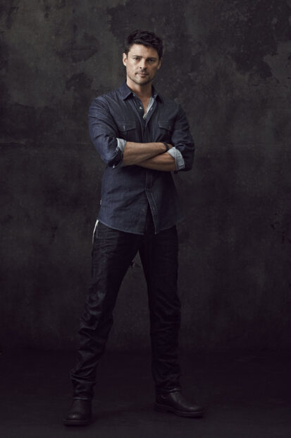 Karl Urban stands with folded arms wearing dark clothing against a textured background, 2K Phone Wallpaper