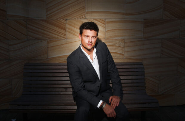Karl Urban wallpapers showing a man in a dark suit sitting on a wooden bench against a patterned wall, 2K Desktop Wallpaper