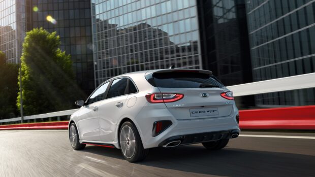 A white Kia hatchback is driving smoothly on an urban road near modern buildings, 4K Desktop Wallpaper