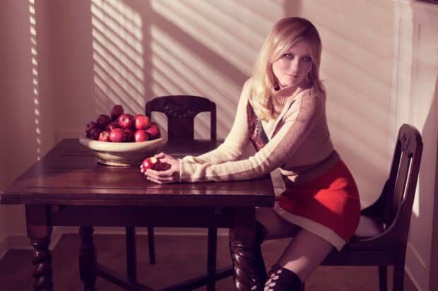 Kirsten Dunst is sitting at a wooden table holding an apple with a bowl of apples beside her, HD Desktop Wallpaper