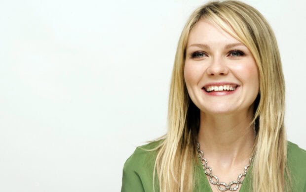 Kirsten Dunst smiles brightly wearing a green blouse and a decorative necklace against a white background, 2K Desktop Wallpaper