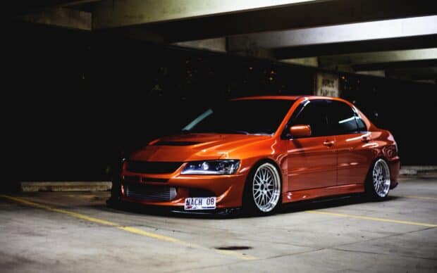 A bright orange Lancer Evo is parked inside a dimly lit parking garage, 2K PC Wallpaper