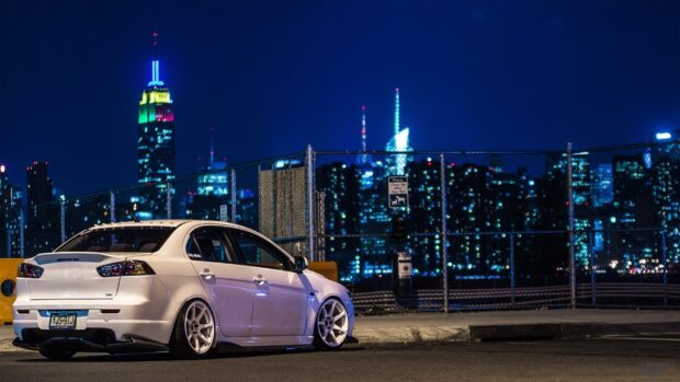 A white Lancer Evo is parked near a fence with city lights in the background, HD Desktop Wallpaper