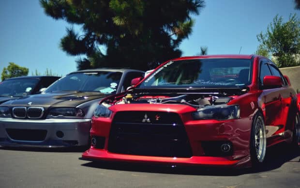 A red Lancer Evo is parked beside other cars in a sunny outdoor setting, HD Desktop Wallpaper