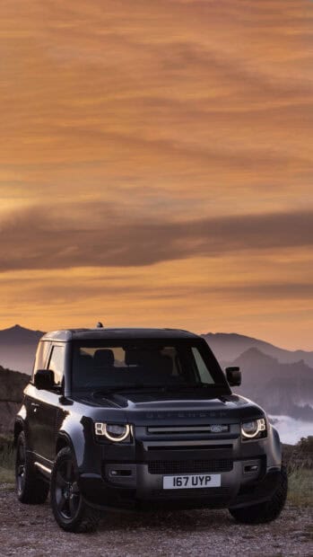 A black Land Rover Defender is parked on a rocky path with mountains and a colorful sunset sky in the background, 2K Mobile Wallpaper