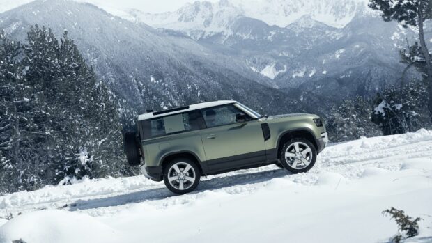 A Land Rover Defender is driving through snow covered terrain with mountain peaks in the background, 4K Desktop Wallpaper