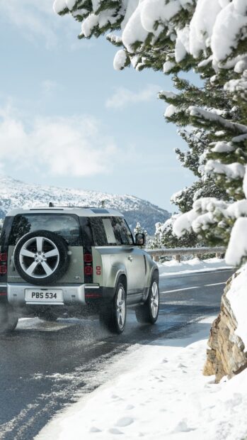 A Land Rover Defender drives along a snow covered mountain road surrounded by pine trees, 4K Phone Wallpaper