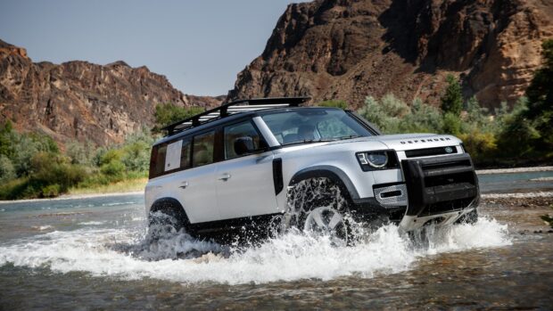 A Land Rover Defender drives through river water with rocky mountains in the background, 4K Desktop Wallpaper