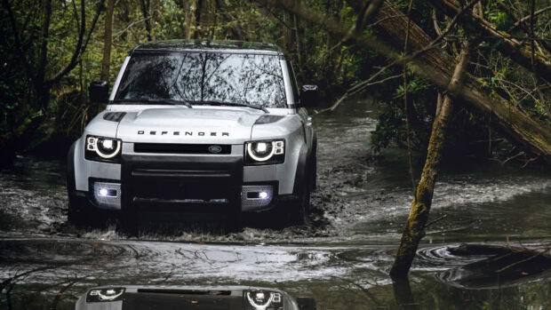 A Land Rover Defender is driving through a muddy forest trail surrounded by trees and water, 4K Desktop Wallpaper
