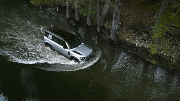 A Land Rover Defender is driving through a shallow river surrounded by trees, 4K Desktop Wallpaper