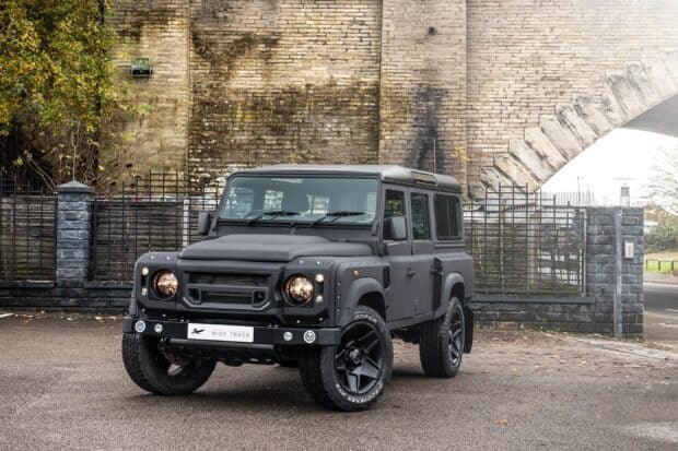 A matte black Land Rover Defender is parked near an old brick wall with greenery around, HD Desktop Wallpaper