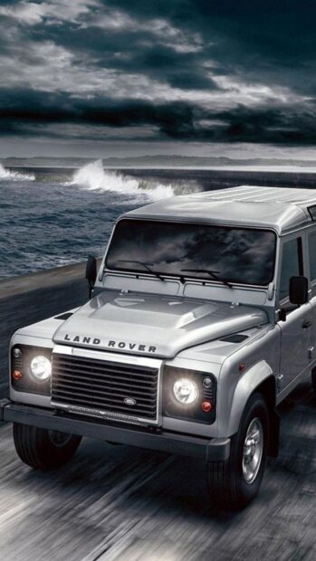 A Land Rover Defender drives along a stormy coastal road with waves crashing nearby, HD Mobile Wallpaper