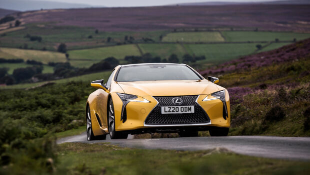 A yellow Lexus LC sports car drives on a rural road surrounded by green fields and hills, 2K Desktop Wallpaper
