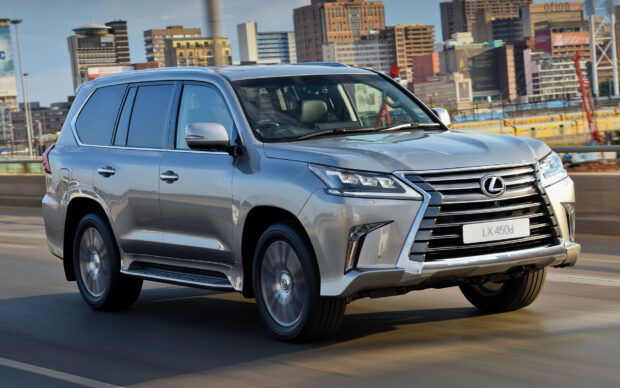 A silver Lexus LX 450d SUV is driving on a highway with city buildings in the background, HD Desktop Wallpaper