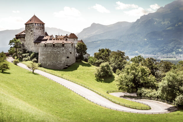 A stone castle overlooks a winding road surrounded by green hills and mountains in Liechtenstein, HD Desktop Wallpaper