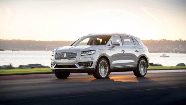 A silver Lincoln Nautilus driving on a waterfront road at sunset with hills in the background, 4K Desktop Wallpaper