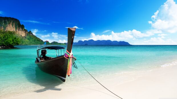 A longtail boat floats near the sandy shore under a bright blue sky in Phuket, 4K Desktop Wallpaper