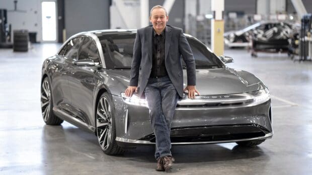 A man stands smiling by a Lucid Motors car in a spacious warehouse, HD Desktop Wallpaper