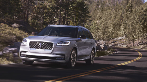 A silver Lincoln car drives along a forest road surrounded by tall trees under clear daylight, 4K Desktop Wallpaper