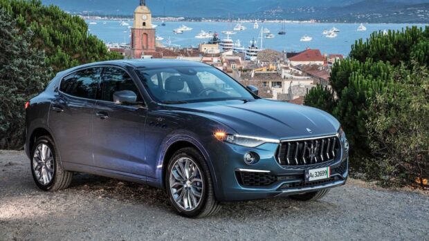 A blue Maserati Levante parked near greenery with a coastal town and boats in the background, HD Desktop Wallpaper