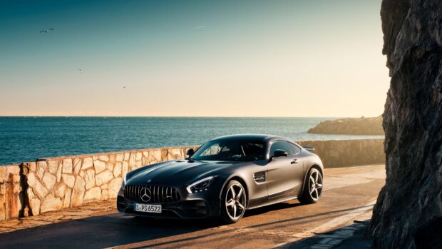 A black Mercedes Benz AMG GT is parked by a stone wall with a calm sea and clear sky in the background, 4K Desktop Wallpaper