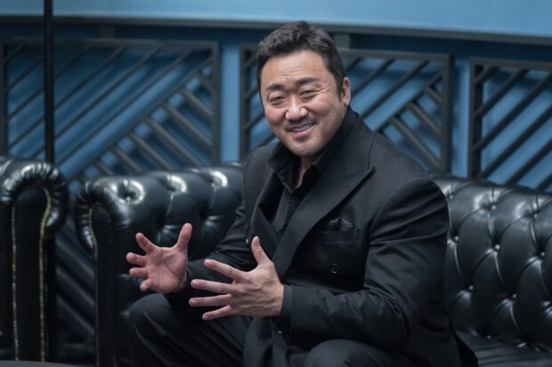Ma Dong Seok smiles warmly while gesturing with his hands on a black leather couch, HD Desktop Wallpaper
