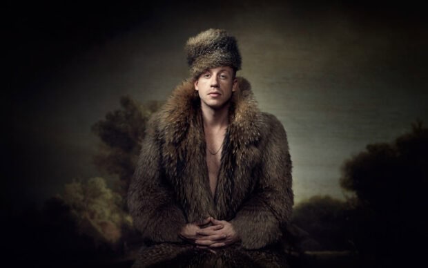 Macklemore is sitting in a fur coat and hat with a blurred nature background, HD Desktop Wallpaper