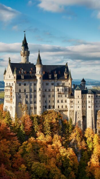 The Neuschwanstein Castle stands surrounded by vibrant autumn trees under a partly cloudy sky, 2K Mobile Wallpaper