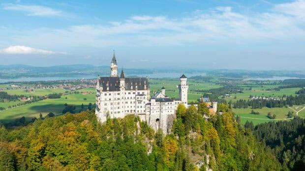 The Neuschwanstein castle is surrounded by lush trees and vast fields under a clear sky, 4K Desktop Wallpaper