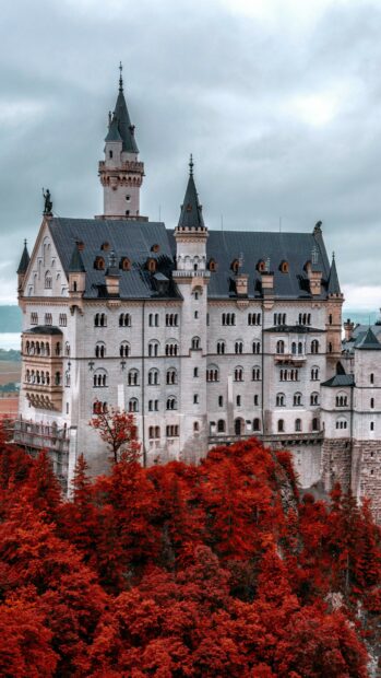 The Neuschwanstein Castle stands prominently surrounded by vibrant red autumn trees under a cloudy sky, 4K Phone Wallpaper