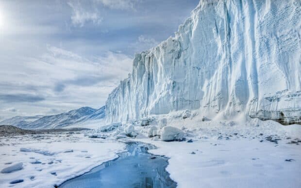A large glacier cliff rises above a frozen snowy landscape with a reflective icy stream, 2K Desktop Wallpaper