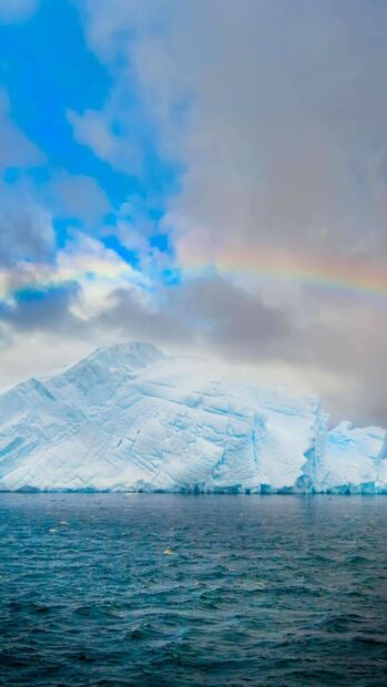 A large glacier is floating in the ocean beneath a cloudy sky with a rainbow, 4K Phone Wallpaper