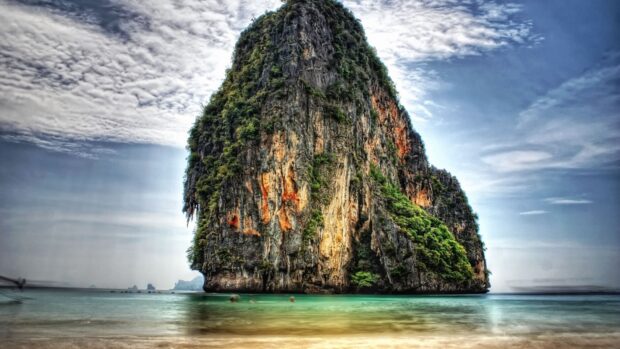 A towering limestone island rises from emerald waters in Vietnam surrounded by sandy beach under a cloudy sky, HD Desktop Wallpaper
