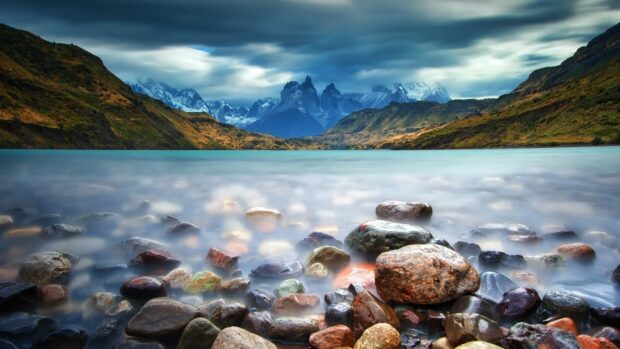A tranquil lake with stones in front of Torres Del Paine National Park mountains under a cloudy sky, HD Desktop Wallpaper