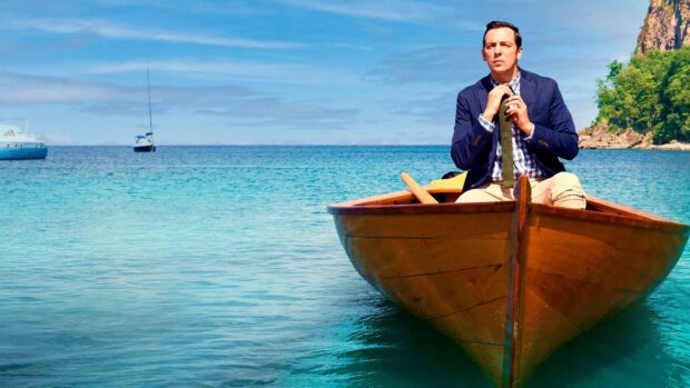 A man adjusts his tie sitting on a wooden boat in a tropical bay from Death In Paradise Tv Series, 4K Desktop Wallpaper