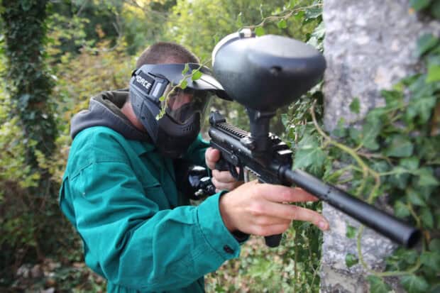 A man wearing protective gear aims a paintball gun behind a stone wall in a forest, HD Desktop Wallpaper
