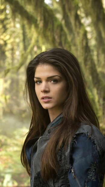 Marie Avgeropoulos stands in a mossy forest wearing a blue jacket with a serious expression, 2K Phone Wallpaper