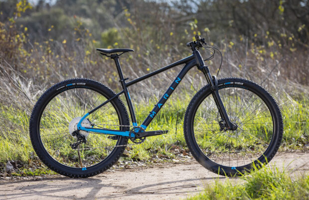 A Marin Bikes mountain bike stands on a dirt trail surrounded by grass and bushes, HD Desktop Wallpaper