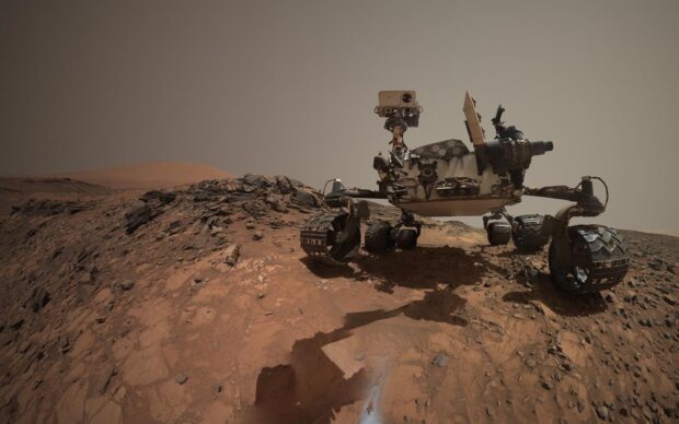 The Mars Curiosity Rover is captured navigating rocky red terrain with distant hills, HD Desktop Wallpaper