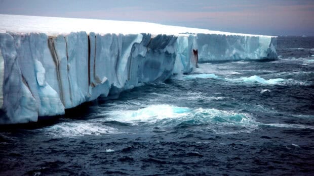 A massive glacier towers over the dark ocean waves under a cloudy sky, 2K Desktop Wallpaper