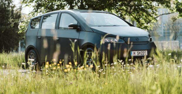 A matte black Sono Motors electric vehicle is parked in a grassy field with trees and buildings behind, HD Desktop Wallpaper