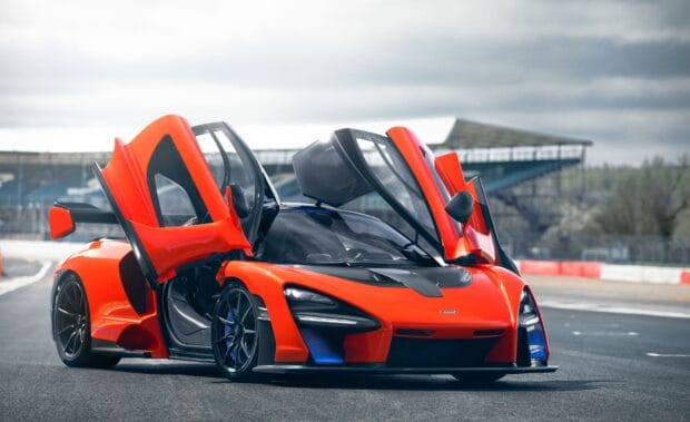 A bright orange McLaren Senna with open doors is parked on a racetrack under a cloudy sky, HD Desktop Wallpaper